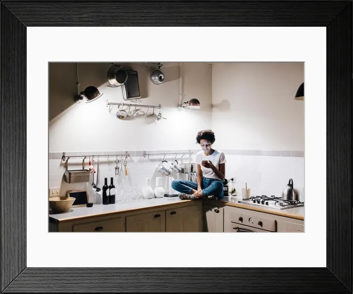 Framed Print : Young woman sitting on kitchen worktop, checking messages on smartphone Framed Print : Young woman sitting on kitchen worktop, checking messages on smartphone