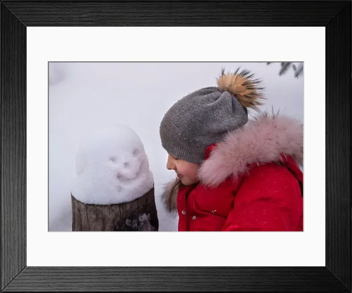 Framed Print : A girl in a winter hat and a red jacket with a small snowman in the snow. The concept of winter fun Framed Print : A girl in a winter hat and a red jacket with a small snowman in the snow. The concept of winter fun
