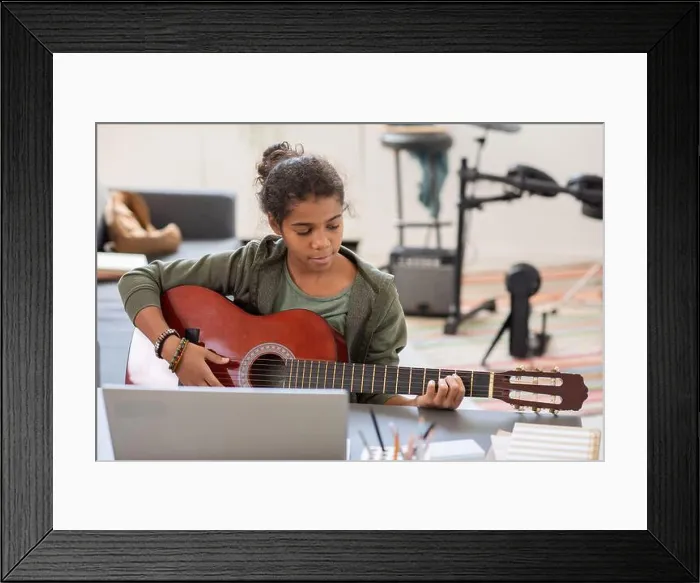 Framed Print : Pretty biracial girl playing acoustic guitar in front of laptop while sitting by table against drumkit in bedroom