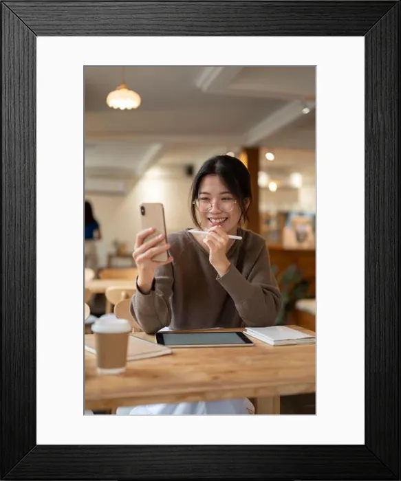 Framed Print : Cheerful young Asian woman in a coffee shop uses her smartphone for a video call and entertainment Framed Print : Cheerful young Asian woman in a coffee shop uses her smartphone for a video call and entertainment