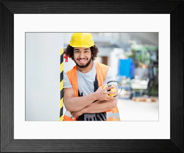 Framed Print : Portrait of confident male worker in protective clothing holding ear protectors at factory Framed Print : Portrait of confident male worker in protective clothing holding ear protectors at factory