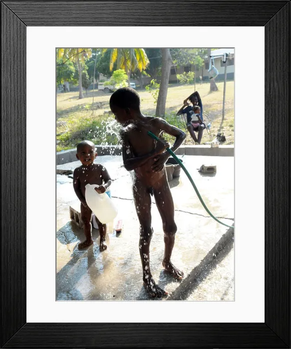 Framed Print : A young black child plays and washes with clean water while a smiling mum looks on Framed Print : A young black child plays and washes with clean water while a smiling mum looks on