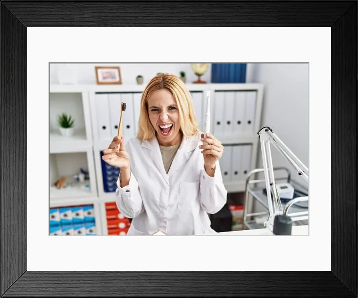 Framed Print : Beautiful blonde dentist woman holding electric toothbrush and normal toothbrush angry and mad screaming frustrated