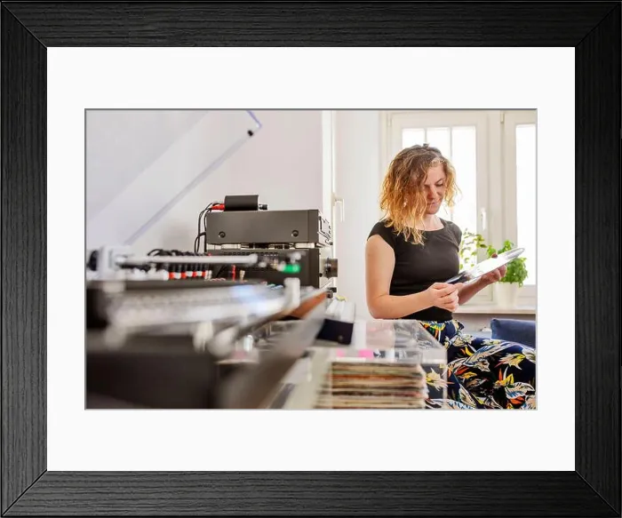 Framed Print : Mid adult woman holding record while sitting by stereo system at home Framed Print : Mid adult woman holding record while sitting by stereo system at home