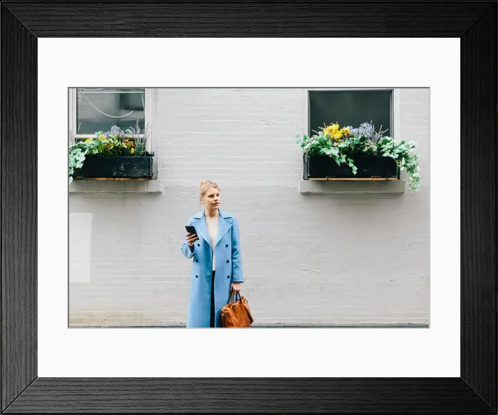 Framed Print : Young woman holding smart phone and purse while standing against building in city Framed Print : Young woman holding smart phone and purse while standing against building in city
