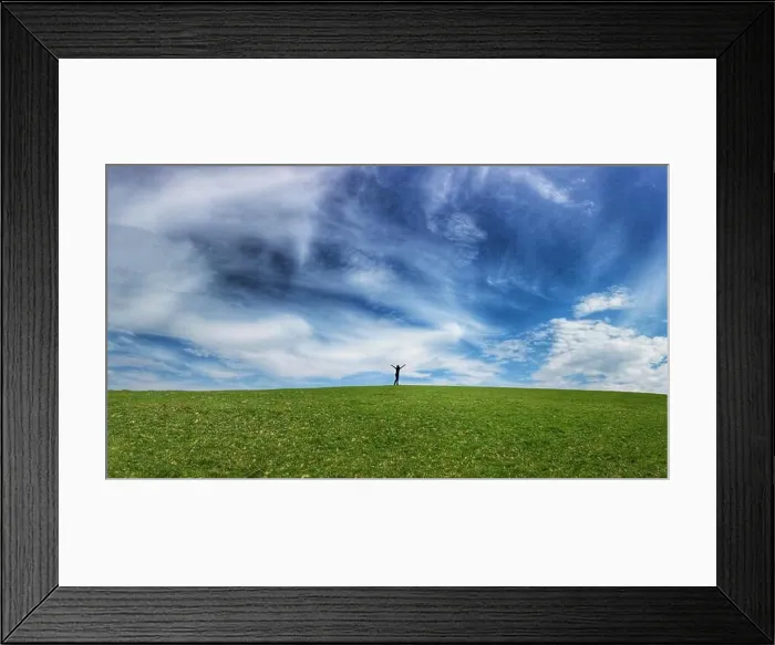 Framed Print : Silhouetted figure with arms held high on horizon of gently rolling grass covered field and blue sky Framed Print : Silhouetted figure with arms held high on horizon of gently rolling grass covered field and blue sky