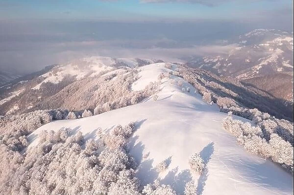 Amazing aerial view of mountains range, meadows and snow-capped peaks in winter time. Forest with frost glowing with bright warm sunrise light