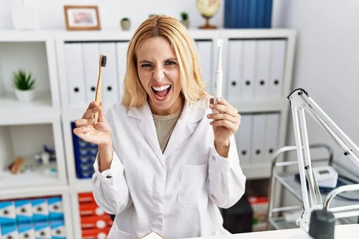 Framed Print : Beautiful blonde dentist woman holding electric toothbrush and normal toothbrush angry and mad screaming frustrated