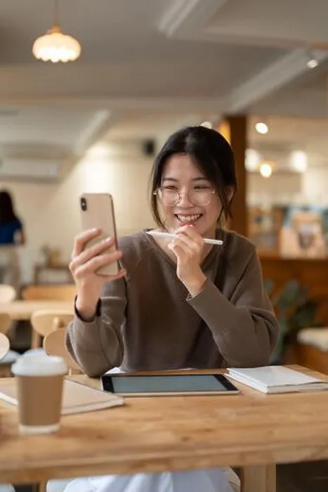 Framed Print : Cheerful young Asian woman in a coffee shop uses her smartphone for a video call and entertainment Framed Print : Cheerful young Asian woman in a coffee shop uses her smartphone for a video call and entertainment