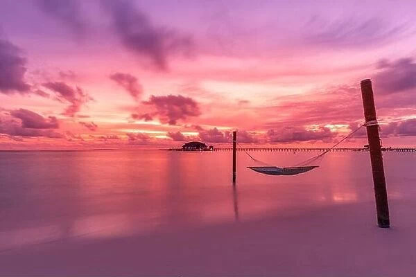 Dramatic sunset sky in tropical island beach. Swing or hammock over water. Purple pink sky clouds, artistic sunrise landscape, coastline