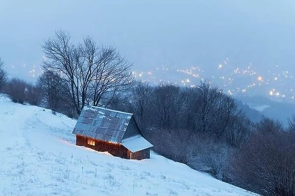 Fantastic winter landscape with glowing wooden house against the backdrop of glowing city lights in fog. Cozy cabin in Carpathian mountains
