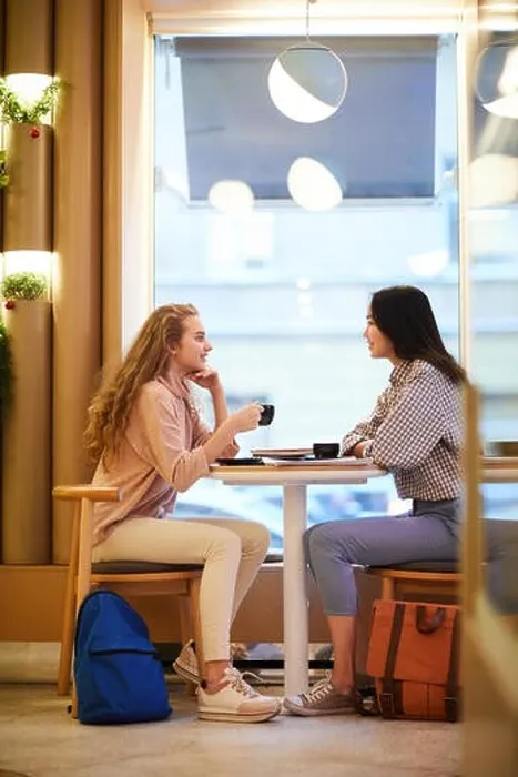 Framed Print : Full length side view portrait of two girls sitting at table in cafe and chatting during break in college, copy space Framed Print : Full length side view portrait of two girls sitting at table in cafe and chatting during break in college, copy space