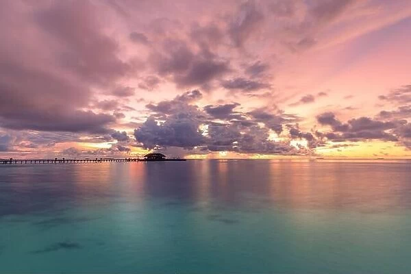 Maldives resort island in sunset with wooden jetty, amazing colorful sky. Perfect sunset beach scenery. Detail of clouds on colorful sky. Vacation