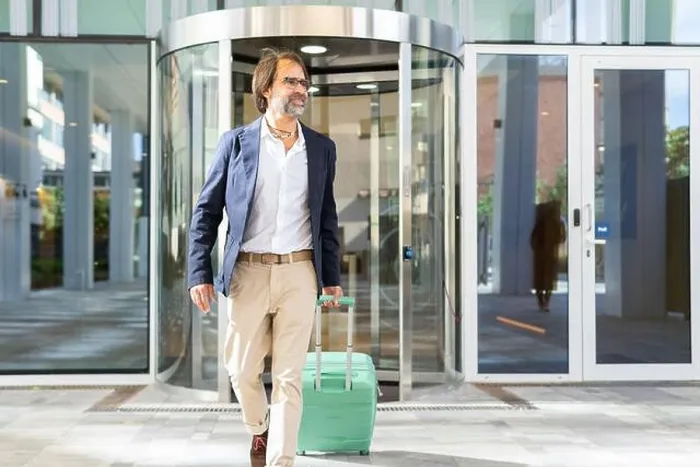 Framed Print : A mature businessman confidently exits a modern glass building, pulling along a light green suitcase and looking away Framed Print : A mature businessman confidently exits a modern glass building, pulling along a light green suitcase and looking away