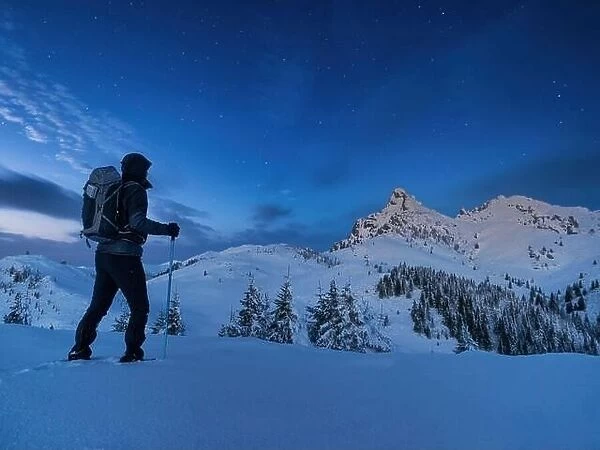 Night photo climber stands on top of a mountain in the snow and looks at the surrounding mountains with starry sky