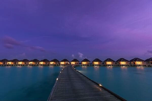 Overwater bungalows in Maldives islands. Tropical paradise island destination, led lights with long jetty under twilight night sky. Exotic landscape