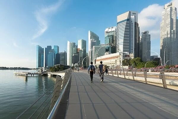 Pedestrians walk along bridge near Marina bay in Singapore with Singapore skyscraper and Merlion park in background