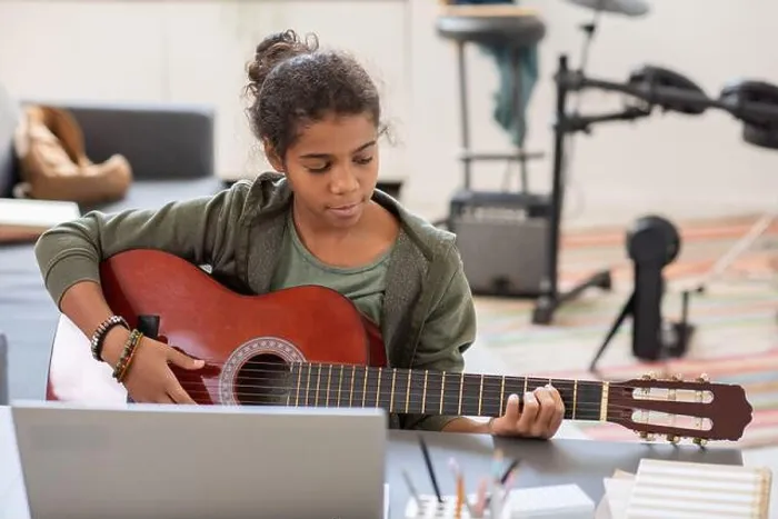 Framed Print : Pretty biracial girl playing acoustic guitar in front of laptop while sitting by table against drumkit in bedroom