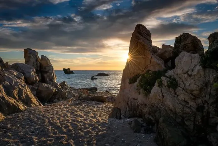 Framed Print : A rocky beach with the sun setting over the sea casting highlights on the sky clouds and sun ray peeping behind a rock Framed Print : A rocky beach with the sun setting over the sea casting highlights on the sky clouds and sun ray peeping behind a rock