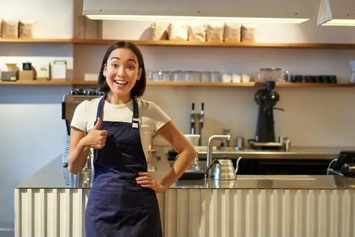 Framed Print : Smiling girl student working part-time in cafe, barista shows thumbs up, wears apron, stands near coffee shop counter Framed Print : Smiling girl student working part-time in cafe, barista shows thumbs up, wears apron, stands near coffee shop counter