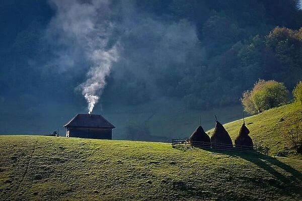 Smoke raising from the chimney of a lonely old wood house near three haystacks on a mountain hill