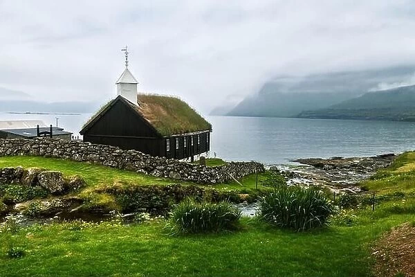 Summer view of traditional turf-top church in faroese village. Beauty landscape with foggy fjord and high mountains