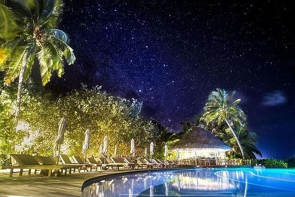 Swimming pool in night illumination under milky way, stars. Palm trees and closed umbrellas with palm trees over infinity swimming pool
