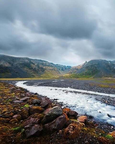 Typical Iceland landscape with river and mountains. Dramatic sky and red stones on a foreground