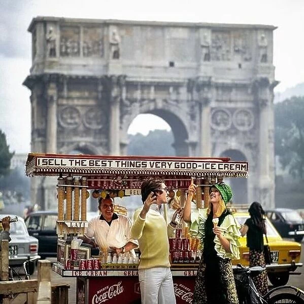 Vintage Rome 1970s, couple having fun by street ice-cream seller in front of triumphal arch of Constantine, Italy, Europe