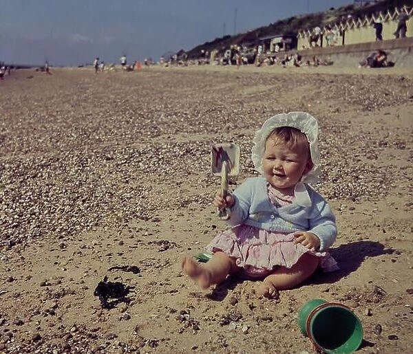 A one year old girl plays with a spade on Clacton Beach in Essex in 1960. The girl wears a white cotton bonnet to protect her from the sun