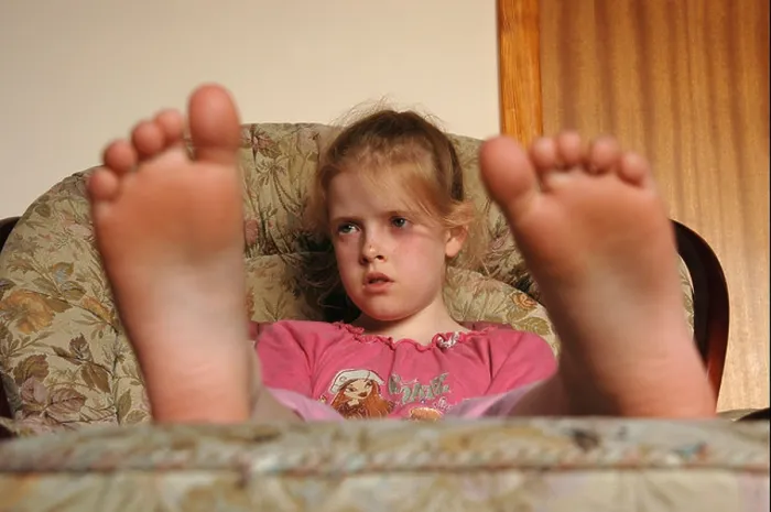 Framed Print : A young seated girl, watches T.V with her bare feet resting on a stool