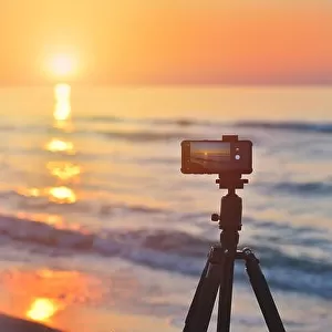 Sunrise over the sea coast. Fireball of the sun above the horizon in a colorful orange sky. Smartphone camera on a tripod in the foreground