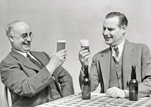 Businessmen Prints: 1930s TWO MEN HAPPY SMILING BUSINESSMEN SITTING AT TABLE HOLDING ADMIRING GLASSES OF BEER EMPTY DARK GLASS BOTTLES ON