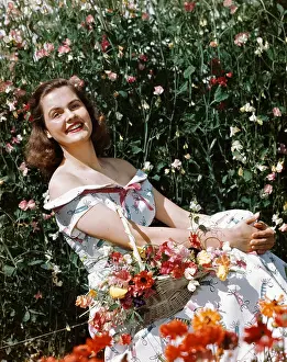 Hars Prints: 1950s SMILING WOMAN SITTING IN MEADOW HOLDING BASKET OF FLOWERS LOOKING AT CAMERA