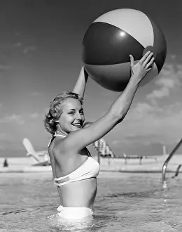 Side Prints: 1950s SIDE VIEW OF BLONDE IN WHITE BIKINI STANDING IN POOL IN WAIST-HIGH WATER HOLDING BEACH BALL IN AIR