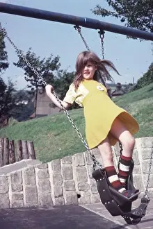 1970s young girl in yellow dress standing up on a swing in a children's playground