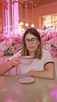 Latina Prints: Adult hispanic woman enjoying coffee in a pink-themed cafe with neon lights and floral decorations