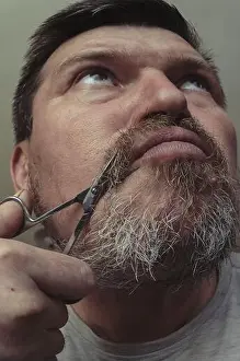 Cheek Prints: An adult man cuts his own beard with small manicure scissors