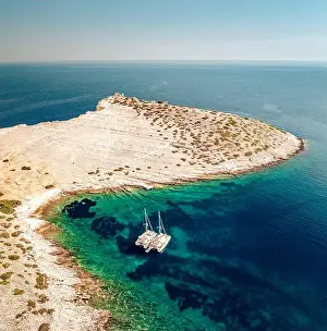 Blue Boat Prints: Aerial view of a catamaran peacefully floats on azure waters near a rocky island