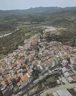 Valencian Prints: Aerial view of a dense cluster of terracotta rooftops and buildings nestled against a backdrop of rolling green hills