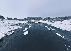 Forest River Prints: An aerial view of a frozen river flowing through snow-covered forests on a cloudy sunset sky background