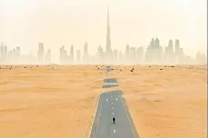 Dubai Skyline Prints: Aerial view of an unidentified person walking on a deserted road covered by sand dunes with the Dubai Skyline
