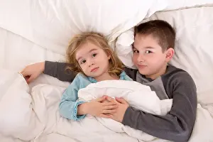 Awake Prints: Affectionate little brother and sister lying in bed cuddling together under the duvet looking up sleepily at the camera