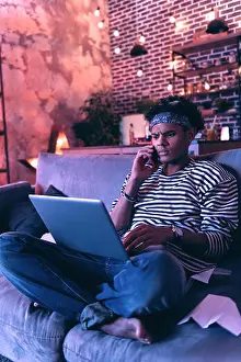 Frown Prints: African American boy sitting barefoot on the couch and checking his mail while talking on the phone