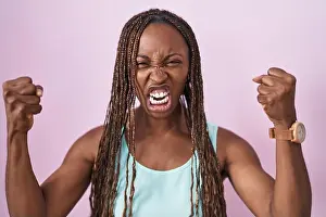 Braids Prints: African american woman standing over pink background angry and mad raising fists frustrated