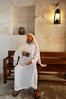 Garb Prints: Arab wearing a traditional white dishdasha robe sitting on a bench with a cup of water