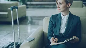 Suitcase Prints: Attractive blonde businesswoman sitting in armchair in hotel lobby and writing in notepad smiling and looking aside