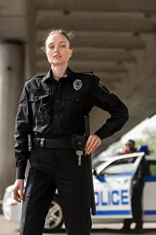 Enforcement Prints: attractive female police officer in uniform looking at camera with blurred partner near car on background