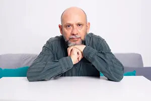 Pants Prints: A bald, well-dressed, bearded man sits thoughtfully on a sofa with his arms resting on a table