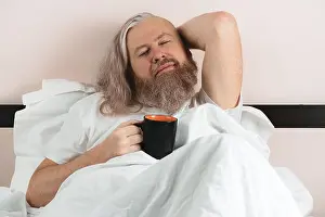 Awakening Prints: Bearded man lying in cozy bed after waking up holding cup of coffee with his eyes closed enjoying